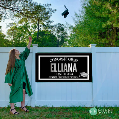 A black and white graduation sign with silver text that reads &#39;Congrats Grad! Elliana Class of 2025 Central Creek High School&#39; with a graduation cap icon. The sign is displayed on a white picket fence.