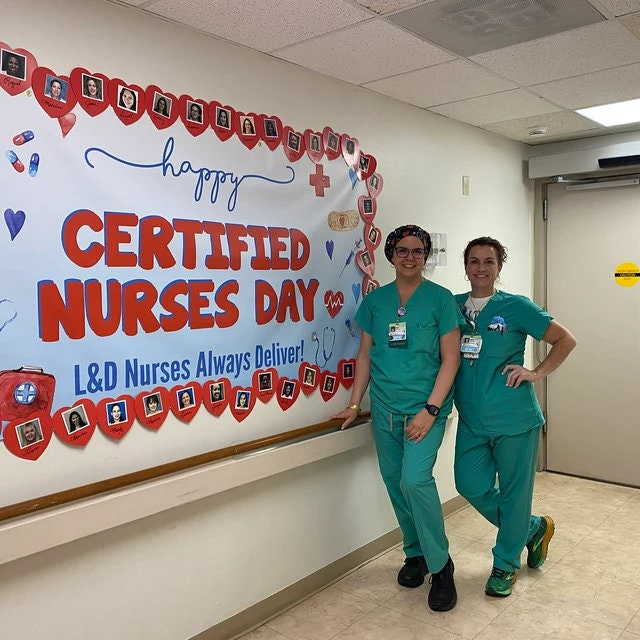 Two nurses in green scrubs stand in front of a white wall with a red and blue sign that says &#39;Happy Certified Nurses Day! L&D Nurses Always Deliver!&#39; The sign is decorated with red hearts and photos of people. The nurses are smiling and looking at the camera.
