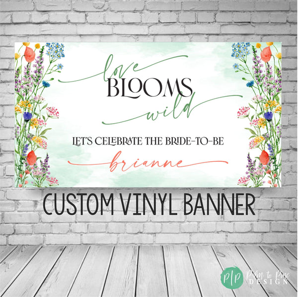 A custom vinyl banner with a watercolor floral design. The banner reads 'love blooms wild' and 'Let's celebrate the bride-to-be' with a personalized name. The banner is green, white, and multicolored flowers with a watercolor effect.