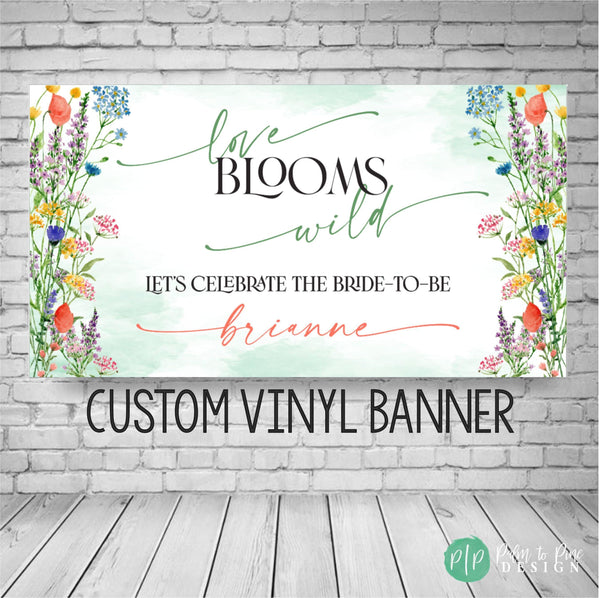 A custom vinyl banner with a watercolor floral design. The banner reads 'love blooms wild' and 'Let's celebrate the bride-to-be' with a personalized name. The banner is green, white, and multicolored flowers with a watercolor effect.